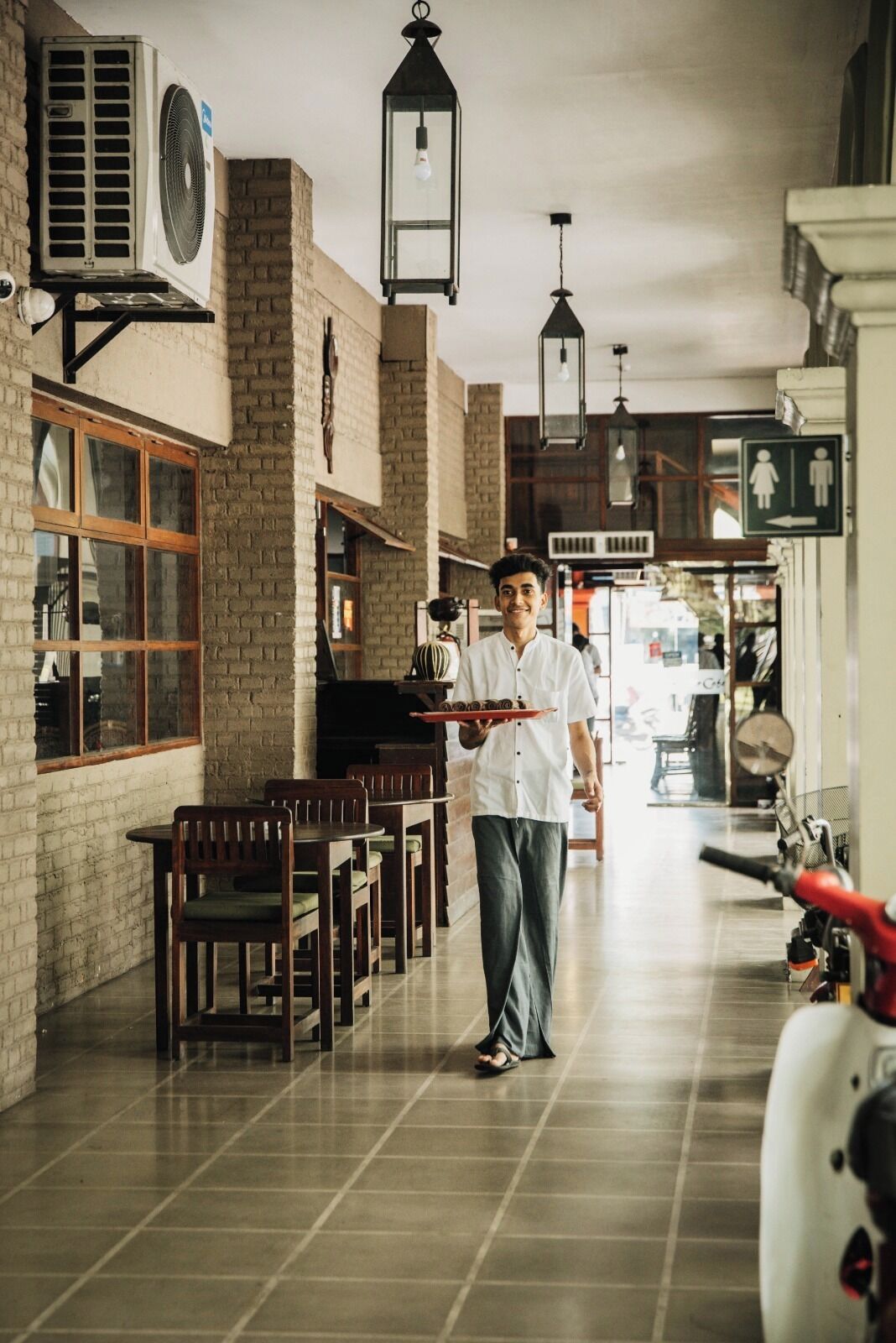 A person carrying a tray with food in a restaurant with brick walls, hanging lanterns, and a tiled floor.