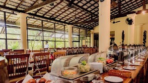 A buffet setup with various food items, wooden tables, and chairs in a spacious room with large windows. - Gimanhala Hotel (Dambulla)