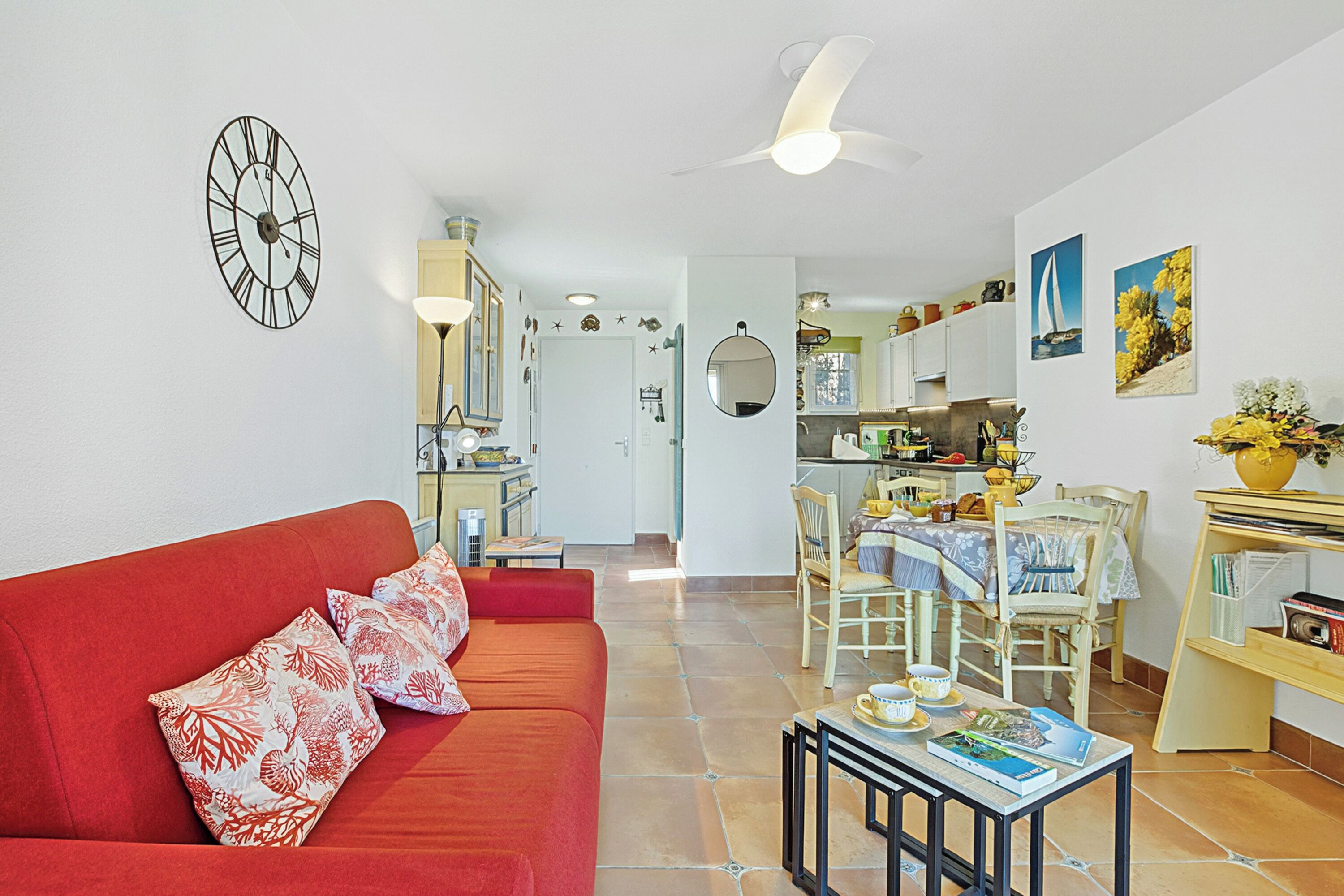 A living room with a red sofa, a dining area with chairs and a table, a kitchen in the background, and a ceiling fan.