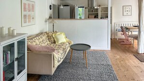 Una sala de estar acogedora con un sofá, una pequeña mesa redonda, un aparador lleno de libros y una cocina al fondo.