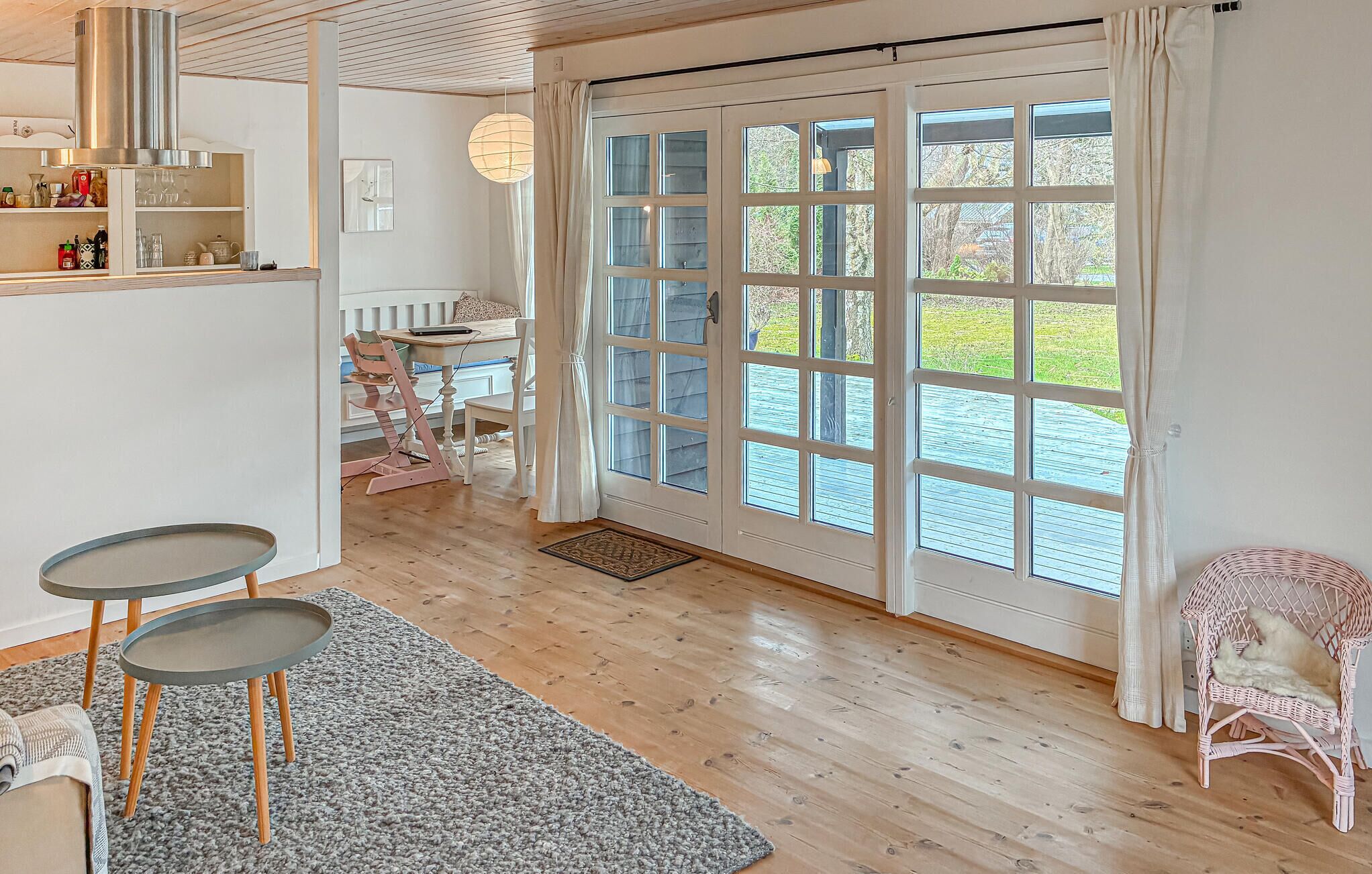 A room with a wooden floor, a dining table with chairs, a wicker chair, a small table with a chair, a large window with curtains, and a door.