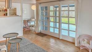 Una habitación con suelo de madera, una mesa de comedor con sillas, una silla de mimbre, una mesita con una silla, un ventanal con cortinas y una puerta.