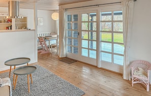 A room with a wooden floor, a dining table with chairs, a wicker chair, a small table with a chair, a large window with curtains, and a door.
