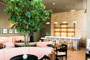 Ein Restaurant mit runden Tischen, gepolsterten Sitzplätzen, Pendelleuchten, einer zentralen Bar und einem großen Innenraum-Baum.