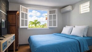 Um quarto com uma cama, uma cômoda de madeira, uma televisão e uma janela com vista para palmeiras.