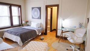 Une chambre à coucher comprenant un lit, un bureau, une chaise, une fenêtre et une porte donnant sur une autre pièce.