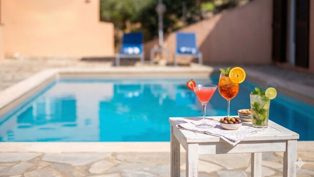 Ein Poolbereich mit Tisch, Getränken, Snacks, einem blauen Sonnenschirm und Liegestühlen im Hintergrund.