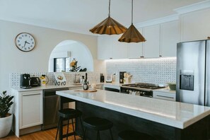 A modern kitchen with a large island, pendant lighting, a clock, and a refrigerator. - Ascot House - Rustic Country Charm, Bowral (Wingecarribee)