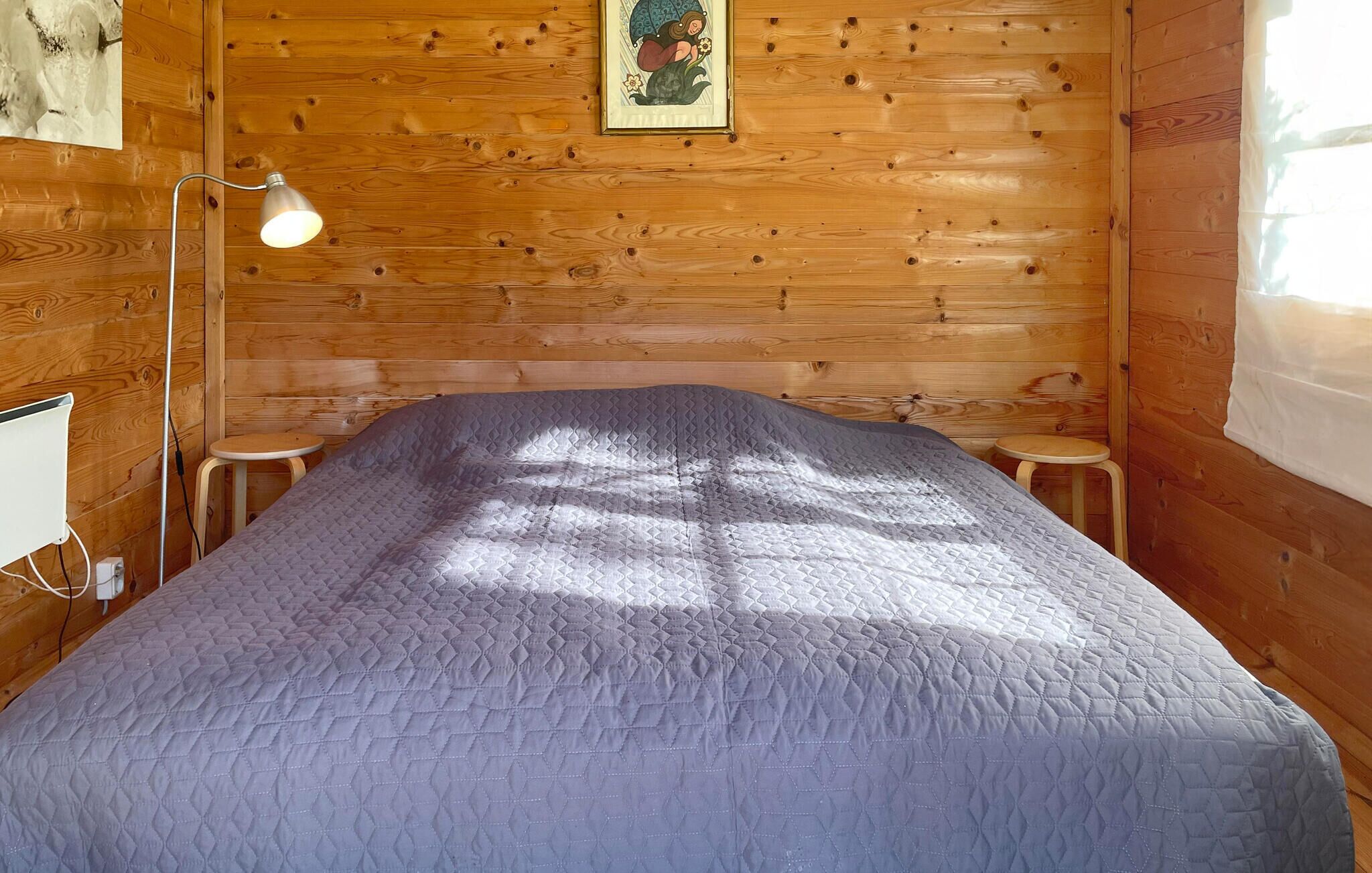 A wooden cabin room with a bed, a desk, a lamp, and a framed picture on the wall.