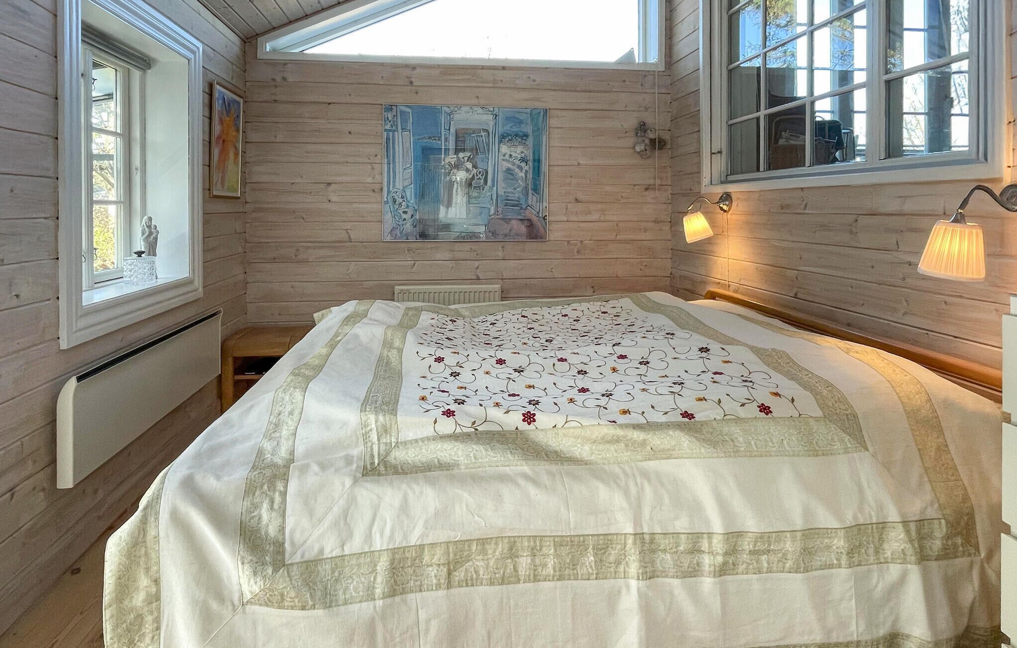 A cozy room with wooden walls, a bed with a floral quilt, a painting on the wall, and a window with a view.