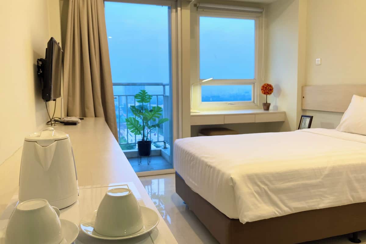 Habitación de hotel con cama, un pequeño balcón con una planta, ventana con vistas y escritorio con tetera y tazas.