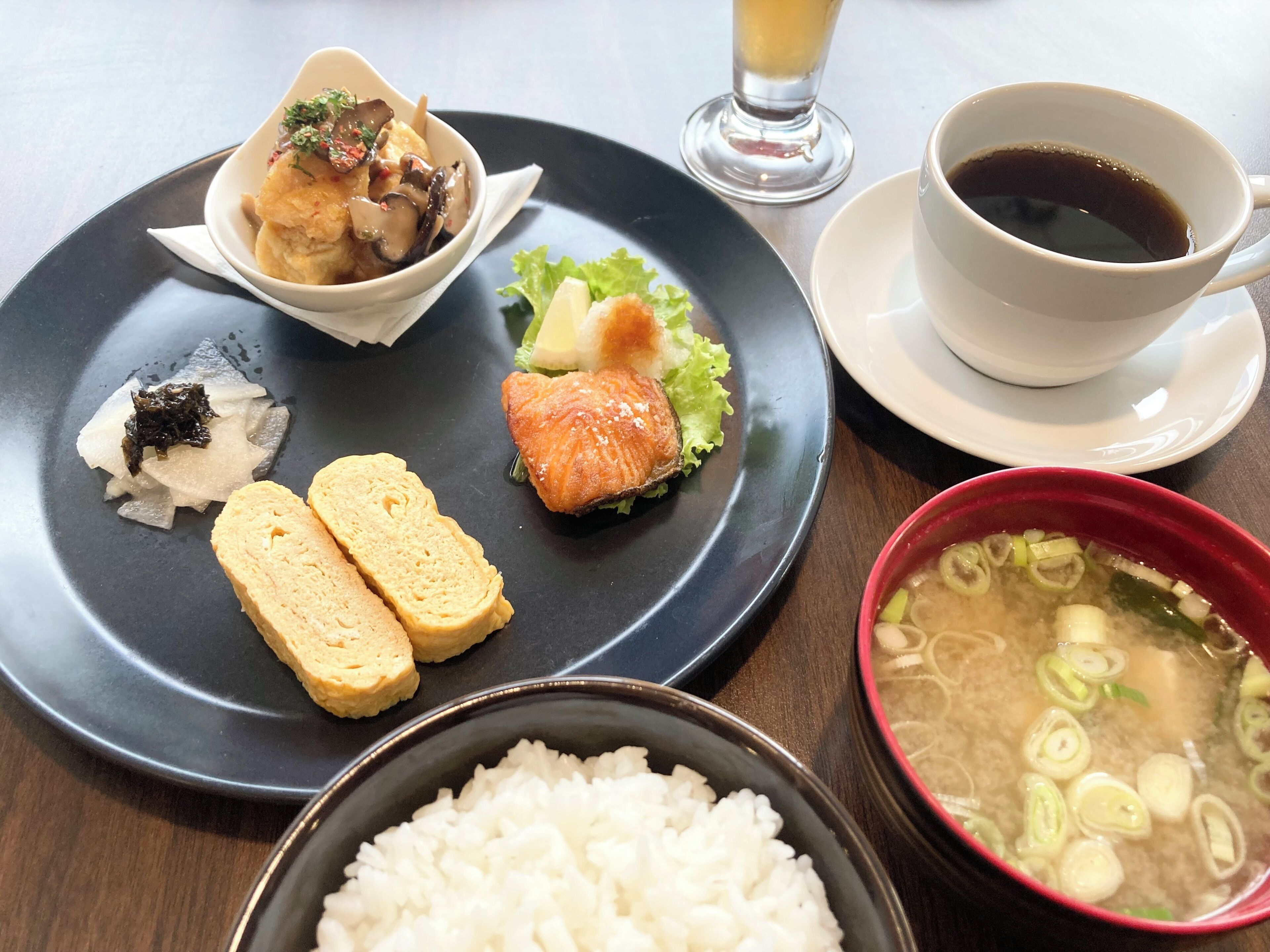 A meal consisting of rice, salmon, tofu, and seaweed on a black plate, accompanied by a cup of tea and a bowl of soup.