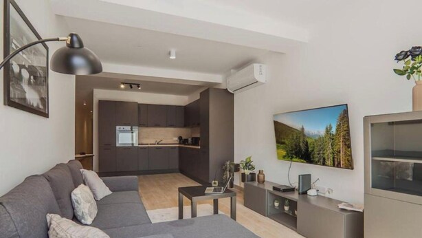 A modern living room with a grey sofa, a coffee table, a TV, and a kitchen in the background. - Ground Floor Maisonette With Communal Pool in Qala (Qala)