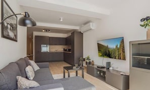 A modern living room with a grey sofa, a coffee table, a TV, and a kitchen in the background. - Ground Floor Maisonette With Communal Pool in Qala (Qala)