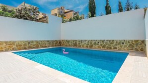 A swimming pool with a pink flamingo float, surrounded by a white wall with a stone border, under a clear blue sky. - Ground Floor Maisonette With Communal Pool in Qala (Qala)