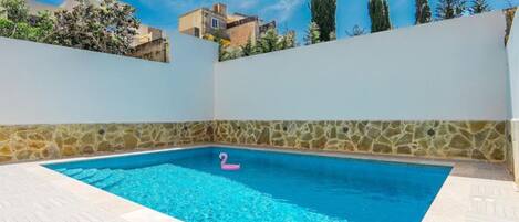 A swimming pool with a pink flamingo float, surrounded by a white wall with a stone border, under a clear blue sky.