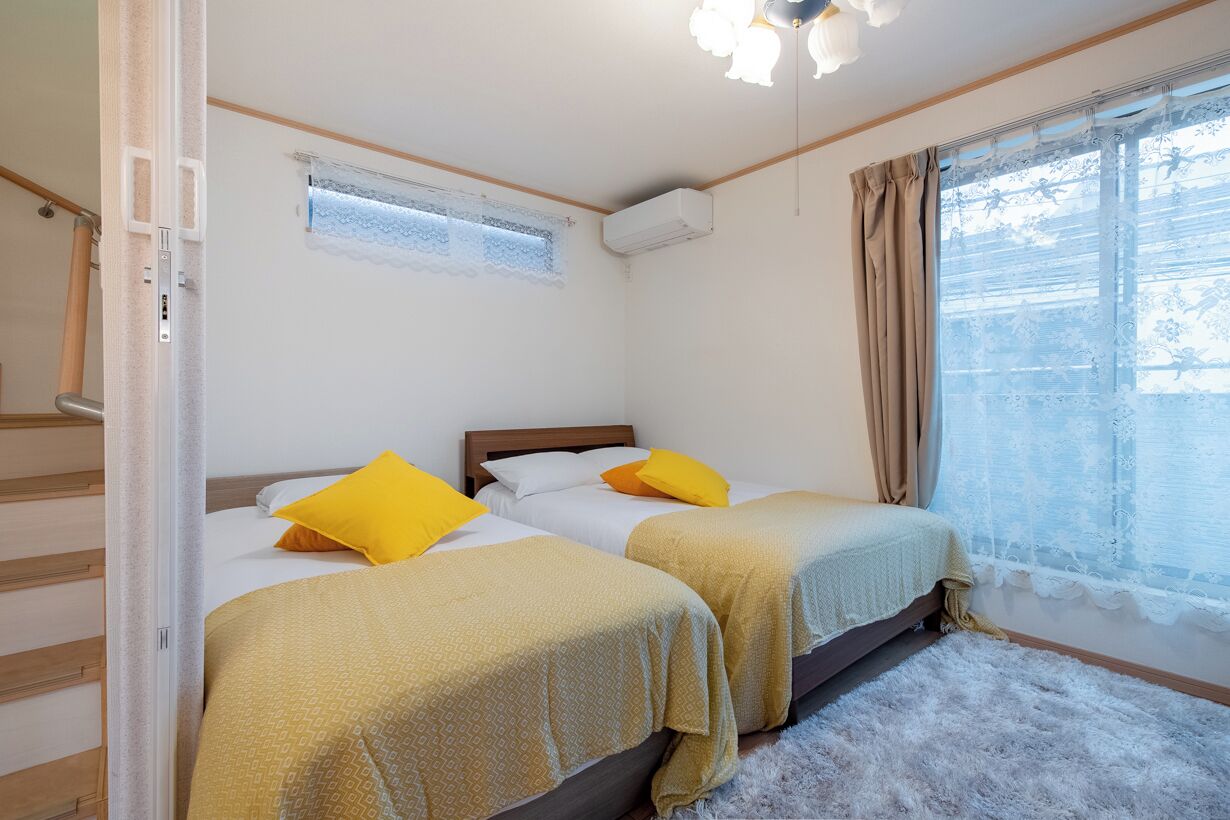 A room with two beds, a window with curtains, an air conditioning unit, and a ceiling light fixture.