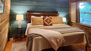 A wooden cabin bedroom with a bed, a nightstand, a lamp, and a window. - Sundance Retreat - Pet Friendly Mountaintop Cabin (Sevierville)