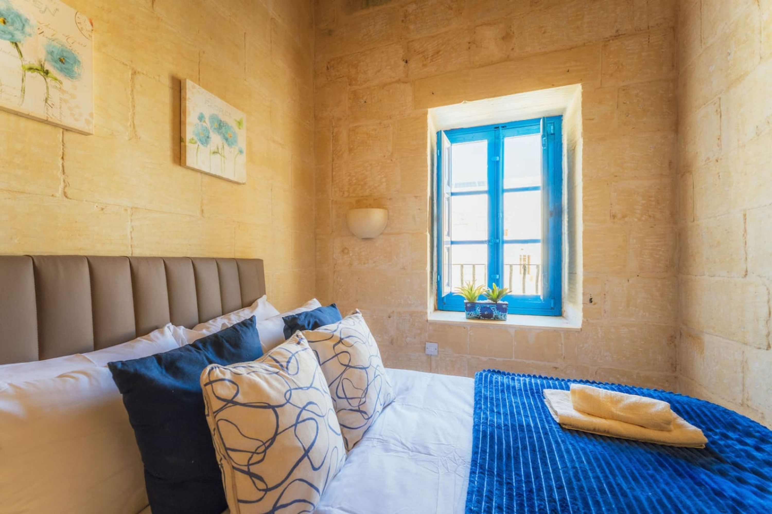 A bedroom with a bed, pillows, a blue blanket, a window with blue shutters, and a wall-mounted bowl.