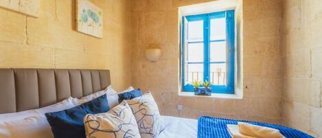 A bedroom with a bed, pillows, a blue blanket, a window with blue shutters, and a wall-mounted bowl.