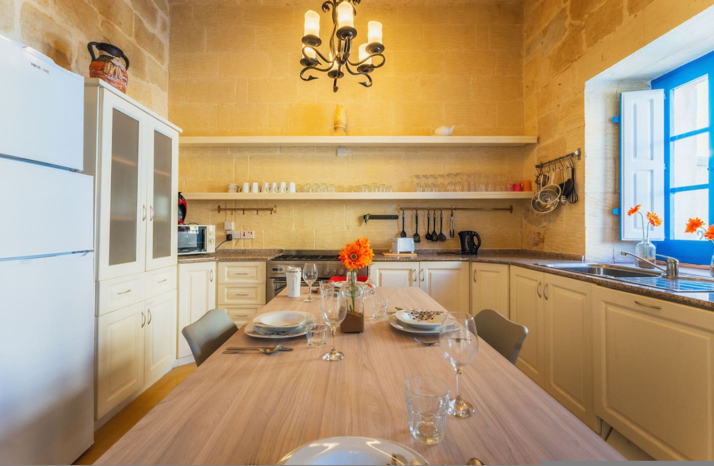 A kitchen with a wooden table set for dining, a chandelier, and a window with blue shutters.