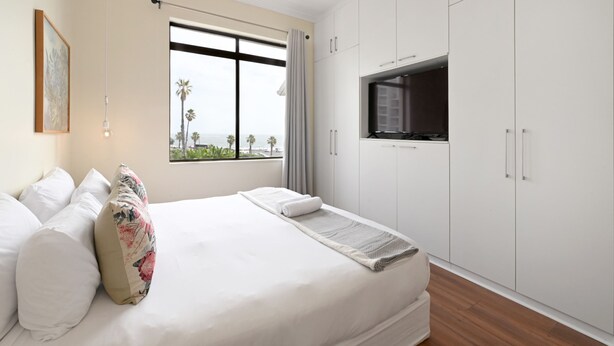 A hotel room with a bed, a TV, white cabinets, and a window with a view. - Sea View Ocean Facing Spacious 1 Bedroom Apartment (Cape Town)