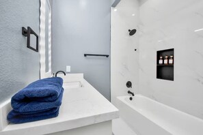 Un baño moderno con un lavamanos de mármol blanco, una toalla azul, una ducha con pared de mármol y un estante pequeño con botellas.