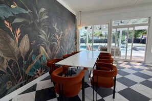 Un comedor con una mesa larga y sillas anaranjadas, un mural con plantas tropicales y un piso a cuadros.