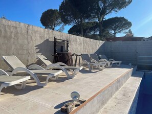 Un área junto a la piscina con sillones de descanso, un muro de concreto y árboles al fondo.