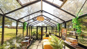 A greenhouse with a yellow sofa, potted plants, and a table with boxes. - Sleeps 30, Private Ranch, 15 acre, Pool, Pond, sauna, pikleball court near DFW (Corsicana)