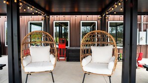 A covered outdoor seating area with wicker chairs and a red chair, wooden walls, and large windows. - Sleeps 30, Private Ranch, 15 acre, Pool, Pond, sauna, pikleball court near DFW (Corsicana)