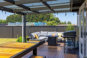 Une terrasse extérieure avec une plateforme en bois, un canapé gris avec des coussins, une petite table et un barbecue.