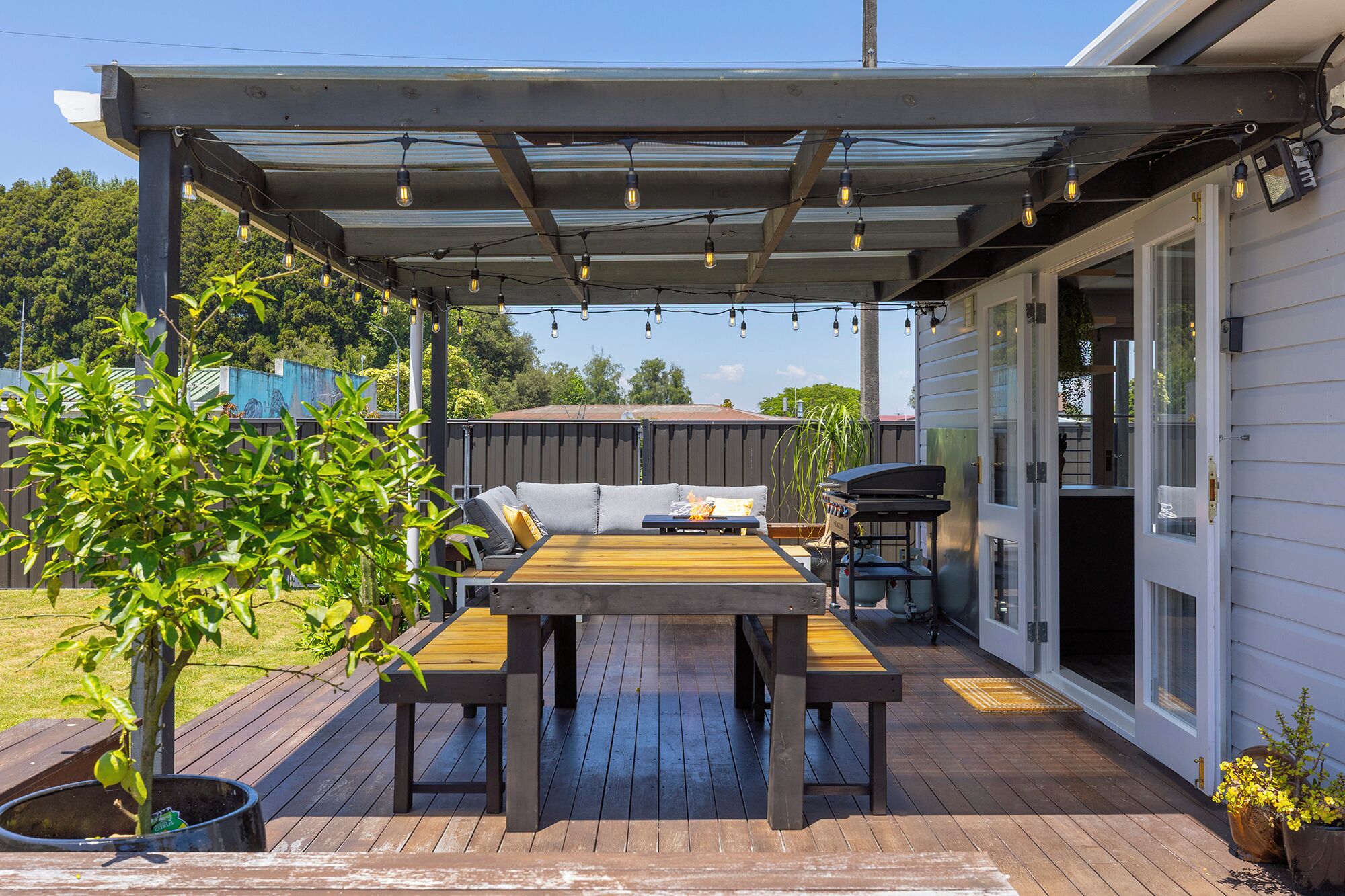 En overdækket terrasse med et træbord og stole, grill og en pergola med lyskæder.
