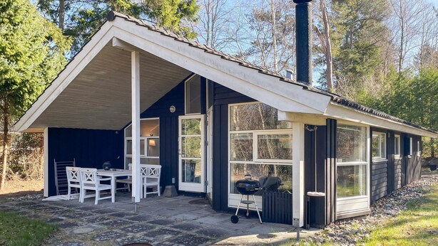 Et lille, etetages hus med en overdækket terrasse, et spiseområde med bord og stole, samt en grill.