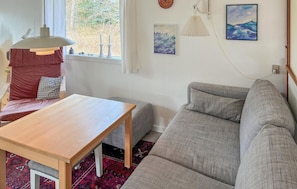 Una sala de estar con un sofá, una mesa de madera, un tapete rojo estampado y un ventanal con vistas a árboles.