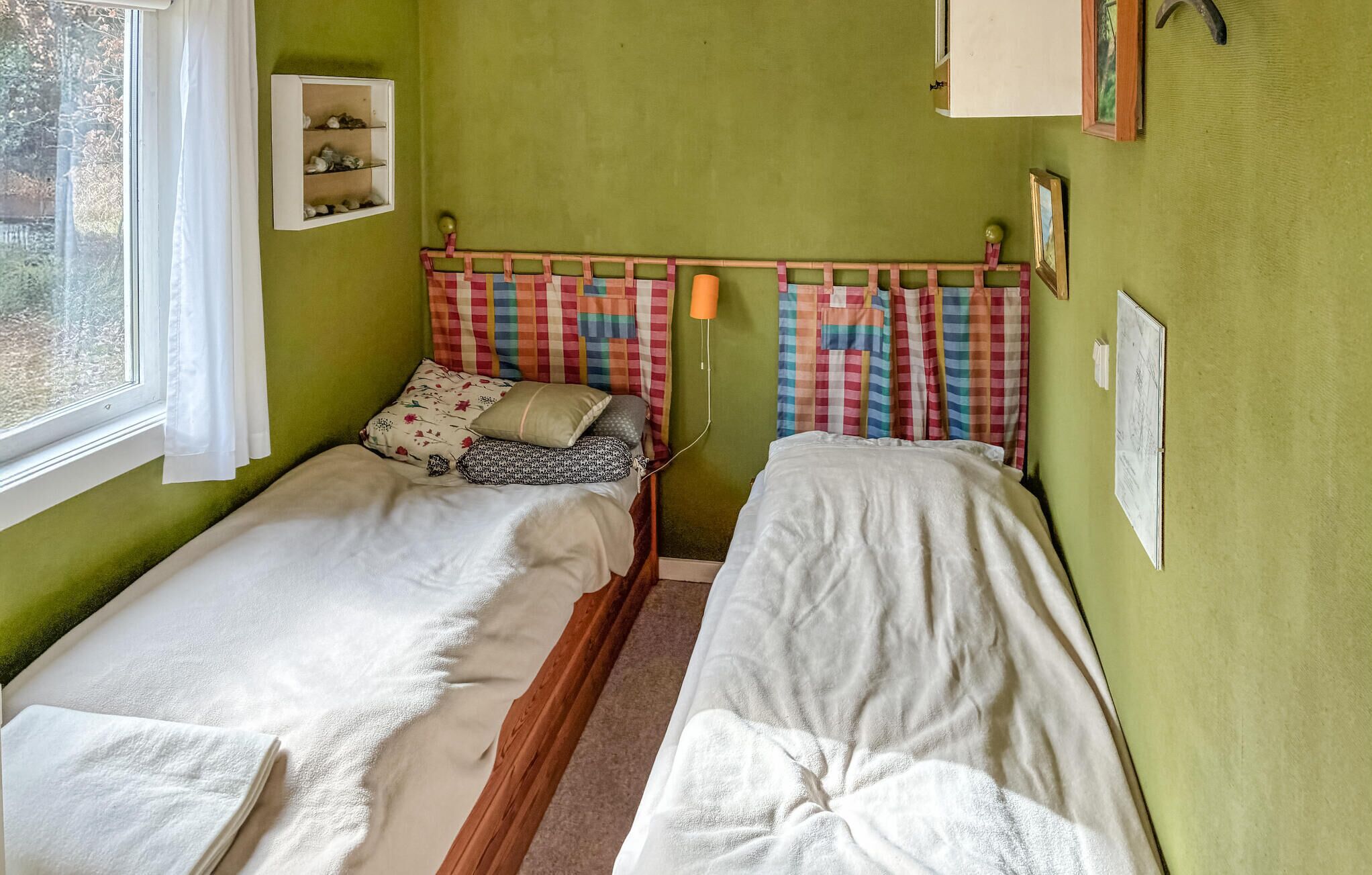 Una habitación con dos camas individuales, una ventana con cortinas blancas, un estante en la pared con vajilla y una lámpara de pared.