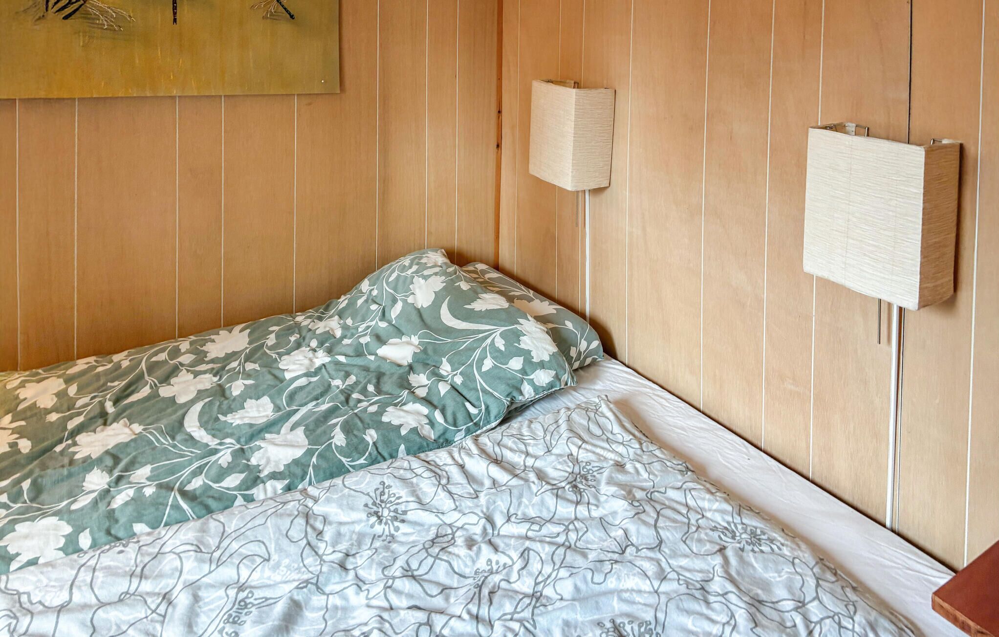 Una cama individual con ropa de cama floral, una lámpara de pared y un panel de madera en la pared.
