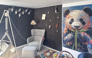 Una habitación con un sillón gris, una hamaca, una lámpara de pared, un cuadro enmarcado y un cuadro abstracto grande.