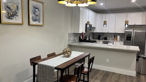 Eine moderne Küche mit weißen Schränken, einem Edelstahl-Kühlschrank, einer Mikrowelle und einer Kaffeemaschine. Ein Esstisch mit Stühlen und einer dekorativen Kugel steht daneben.