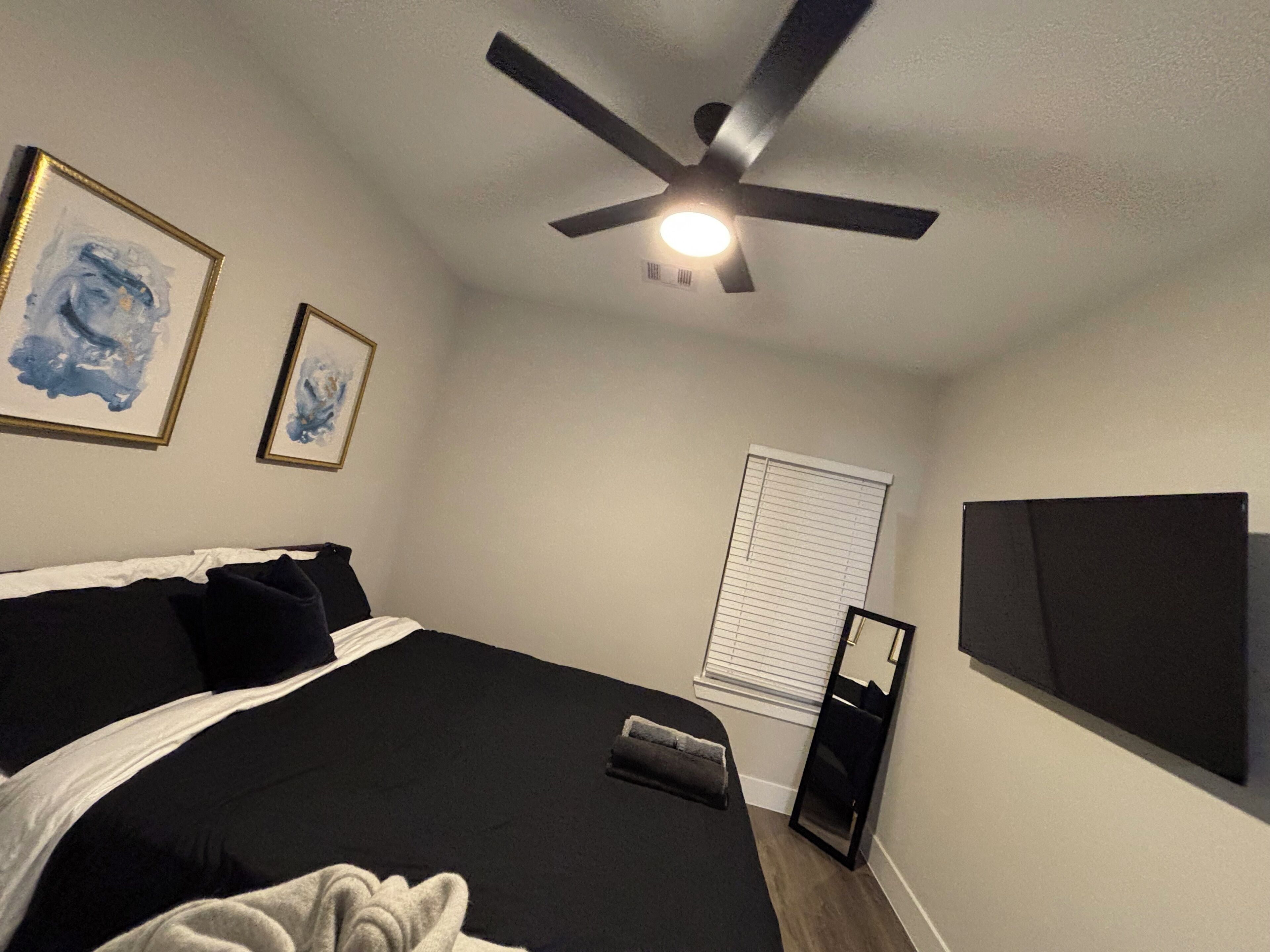 Une chambre avec un lit, un ventilateur de plafond, des œuvres d’art accrochées au mur, une fenêtre avec des stores et un haut-parleur noir.