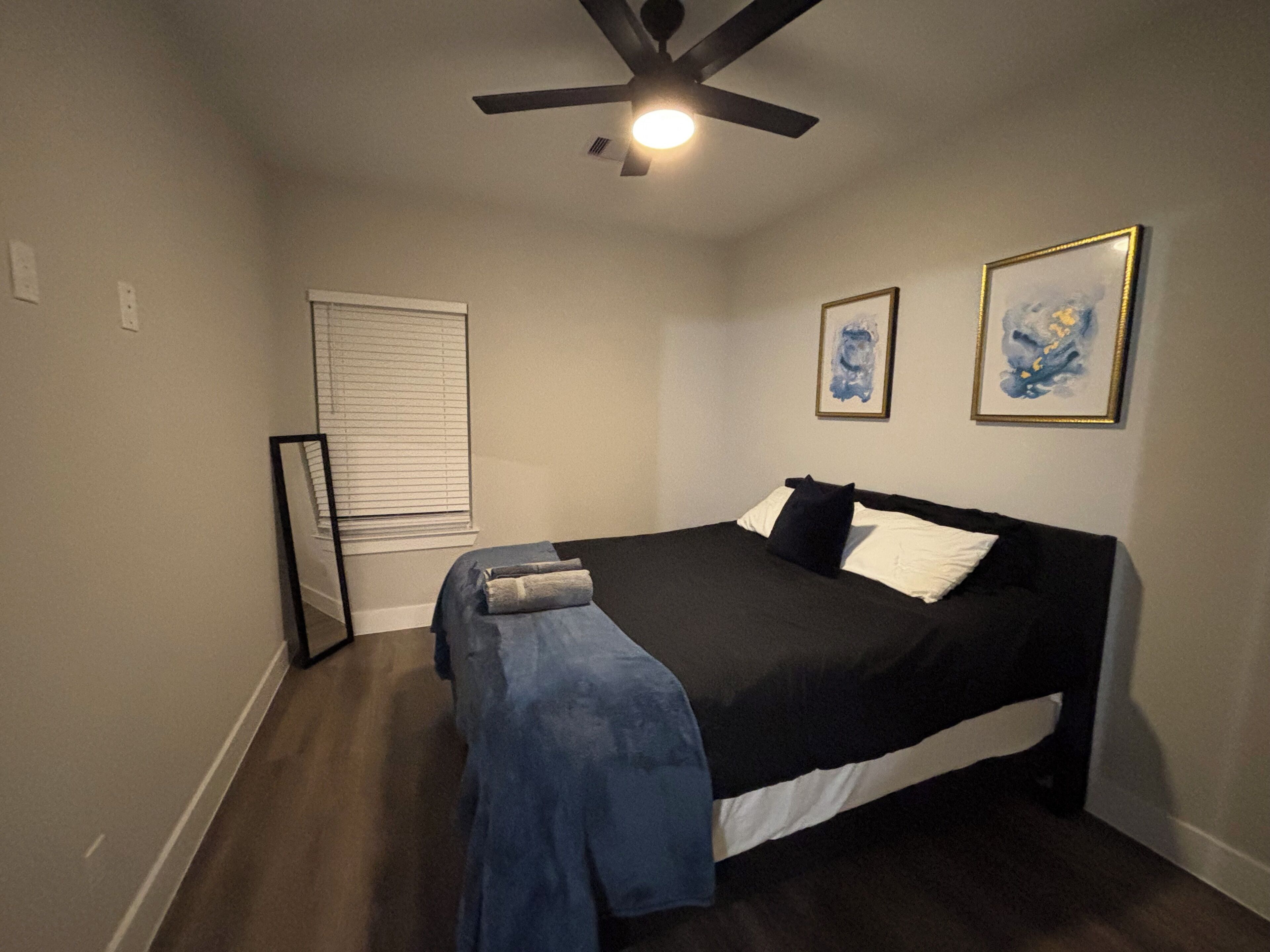 Een slaapkamer met een bed, een plafondventilator, een raam met jaloezieën en twee ingelijste kunstwerken aan de wand.