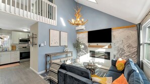 A modern living room with a sofa, a chandelier, and a flat-screen TV mounted on the wall. - Hot Tub, Jungle Gym, Fire Pit - Private Cabin Gem! (Tobyhanna)