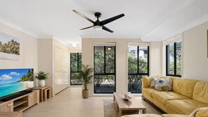 A spacious living room with a large flat-screen TV, a yellow leather sofa, a ceiling fan, and large windows offering a view of greenery. - Hawks Nest Hideaway | Spa, BBQ, Bikes & Pool Table (Hawks Nest)