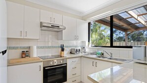 A modern kitchen with white cabinetry, a built-in oven, a stainless steel range hood, and a window overlooking greenery. - Hawks Nest Hideaway | Spa, BBQ, Bikes & Pool Table (Hawks Nest)