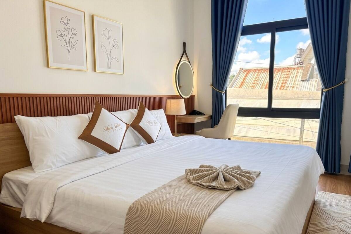 Una cama impecablemente tendida con sábanas blancas y una manta de espiga, una mesita de noche con lámpara, y vistas de la ciudad a través de amplios ventanales.
