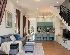 A modern living room with a sofa, ottoman, and a dining area with chairs and a kitchen in the background. - Minh Duc Villa (Lam Vien - Da Lat)