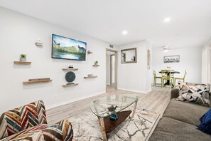 A modern living room with a glass coffee table, a sofa with patterned cushions, a dining area with a green chair, and a painting on the wall. - Charming 2 BR near Deep Ellum (Dallas)