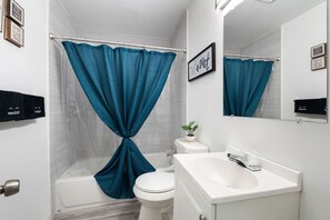 A bathroom with a bathtub, toilet, and sink, featuring teal curtains and a 'Take a seat' sign. - Charming 2 BR near Deep Ellum (Dallas)