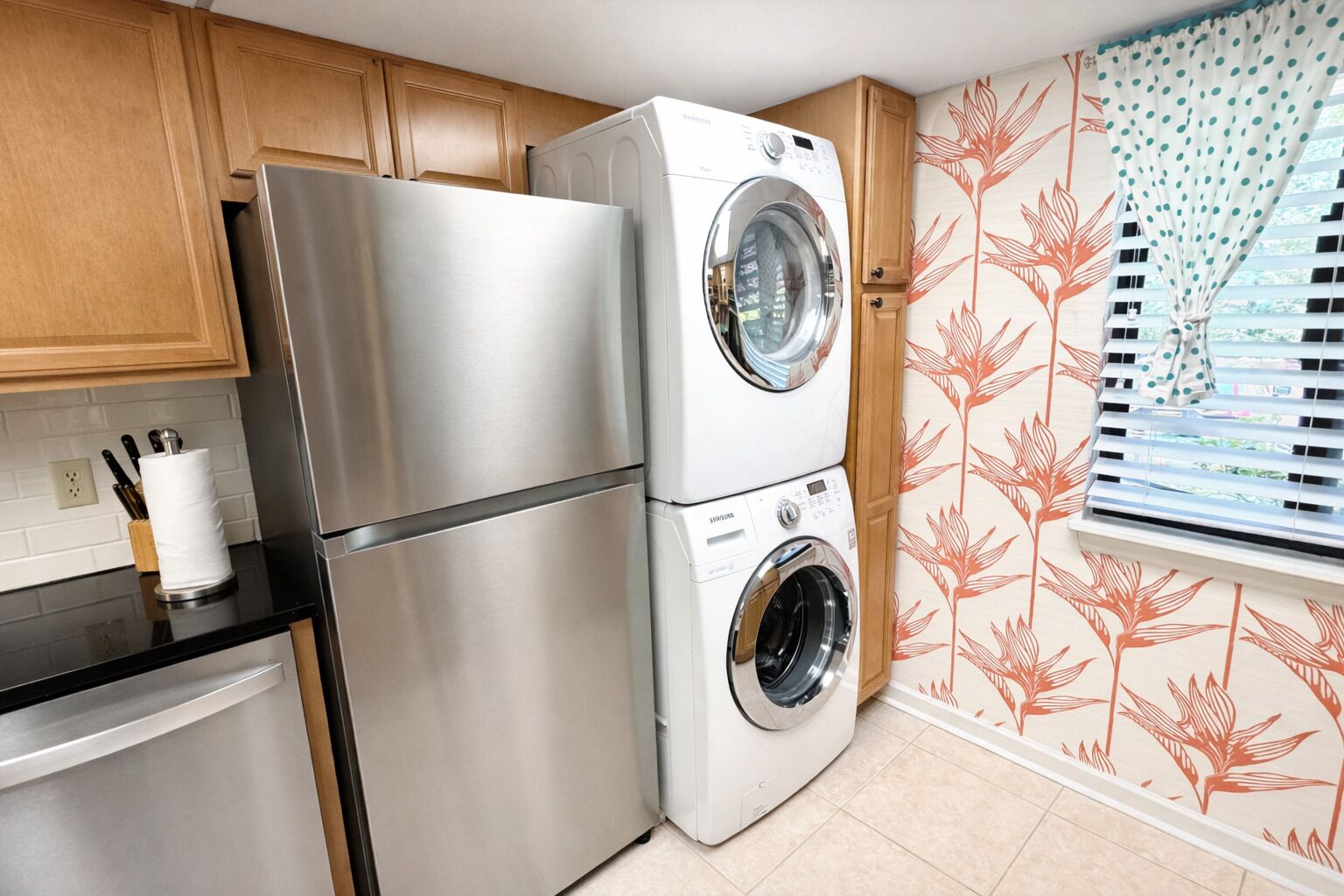 Cocina compacta con refrigerador de acero inoxidable, lavadora y secadora integradas, gabinetes de madera y una ventana con persianas.