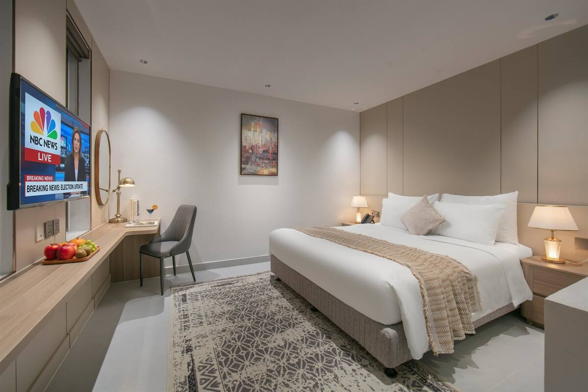 Une chambre d’hôtel comprenant un lit, une table de chevet, une lampe, une chaise, un téléviseur diffusant les informations de NBC News et un plateau de fruits.